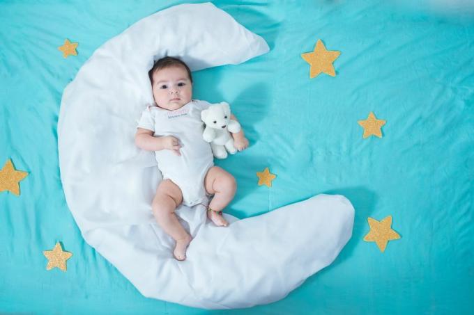 graži lotynų mergaitė, dviejų mėnesių, guli ant balto mėnulio formos paklodės su geltonomis žvaigždėmis abiejose pusėse ir mėlynu paklode po juo