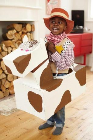 mažas berniukas, apsirengęs kaubojumi su kaubojaus kepure, languotais marškiniais ir bandana su kartoniniu arkliu per juosmenį