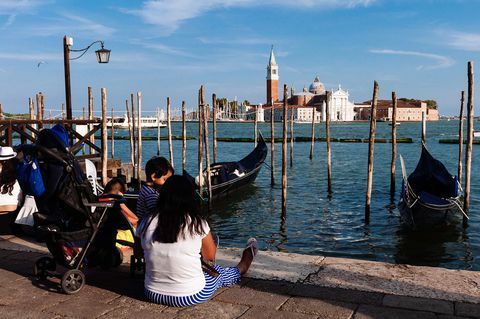 Turistai sėdi ant žemės Venecijoje žvelgdami į San Giorgio bažnyčią