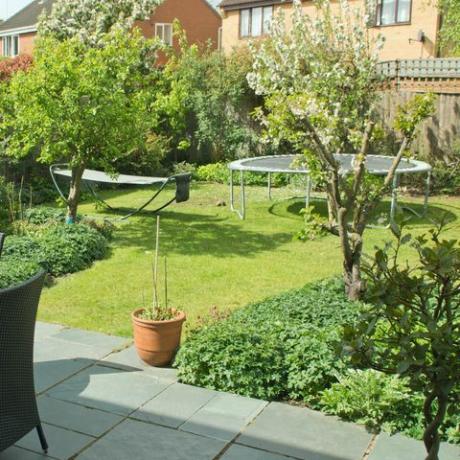 užpakalinis sodas Anglijos priemiesčio nuosavybėje su kėdėmis ir stalu ant šiferio terasos ir batuto bei hamako ant pievelės fone