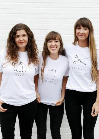trys moterys baltais marškinėliais, ant marškinėlių užrašytos ir pavaizduotos uolos, popierius ar žirklės
