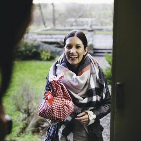 moteris šypsosi stovėdama ant durų laiptelio, aplankydama draugą, ji yra apsivilkusi šiltais drabužiais ir laiko dovaną, supakuotą į raudonai baltą languotą audinį