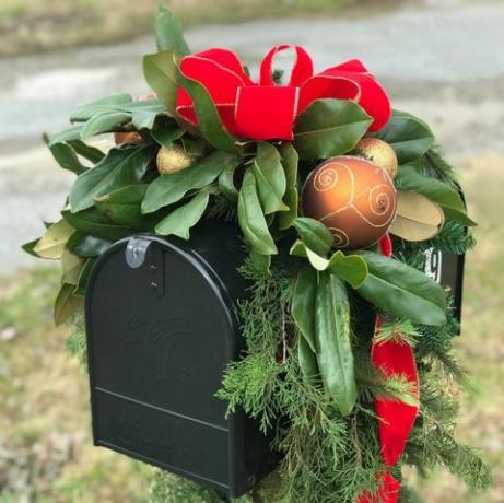 juoda pašto dėžutė, dekoruota šventėms, auksiniais kalėdiniais kamuoliukais, raudona juostele ir daugybe žalumos, įskaitant magnoliją ir kedro lapus