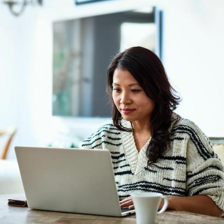 30 metų amžiaus vidurio suaugusi moteris sėdi prie stalo ir žiūri į kompiuterį, dirba nuotoliniu būdu, laisvai samdoma darbuotoja, smulkus verslas