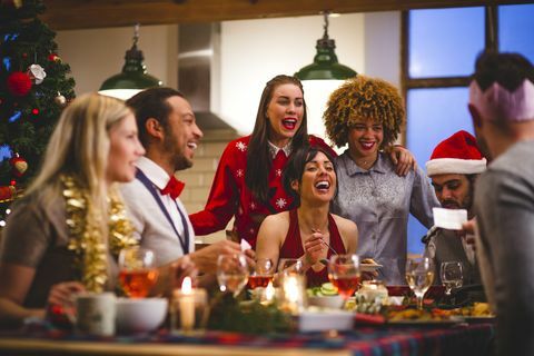 grupė draugų juokiasi sėdėdami prie stalo valgydami ir geriantys per Kalėdas vienas vyras skaito pokštą iš kalėdinio krekerio vakarėlio kepuraitės ir kalėdiniai megztiniai dėvimi